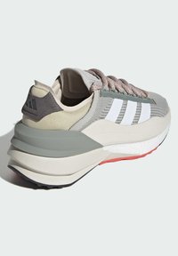 Zapatilla deportiva de color gris claro con parte superior de malla, tres rayas blancas, cuello acolchado y suela de goma contrastante con acentos en rojo y negro.