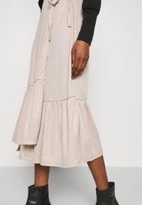 Gros plan sur une jupe midi beige à volants avec une ceinture à nouer, portée avec un haut noir à manches longues et des bottines noires.