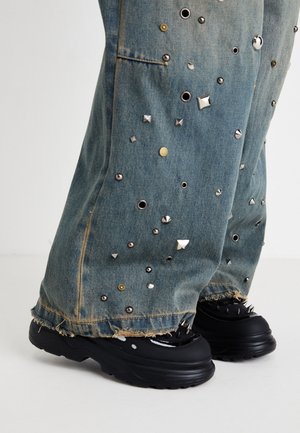 Pantalon en denim évasé avec des clous et œillets métalliques. Ourlet effiloché et poches visibles. Porté avec des chaussures noires chunky présentant des pointes.