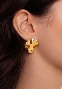 Goldene Metallohrringe mit organischer Form, die eine glatte, reflektierende Oberfläche und leicht strukturierte Kanten aufweisen, getragen am Ohrläppchen.