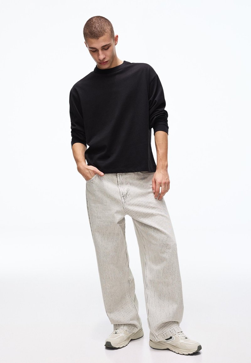 Jeune homme portant une chemise noire à manches longues, un pantalon beige à larges rayures et des baskets blanches, debout avec une main dans la poche.