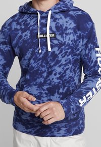 Hoodie tie-dye marine avec des cordons blancs et une poche avant. Présente le logo "HOLLISTER" sur la poitrine et la manche en blanc contrastant. Tissu doux.