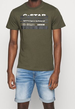 Olivengrøn T-shirt med fed hvid og sort tekstprint. Rund halsudskæring, korte ærmer. Bæres med lyseblå rullede denimshorts.
