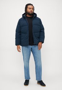 Navy puffer jacket with a hood, featuring quilted design and front pockets, worn over a black shirt and paired with blue jeans and black sneakers.