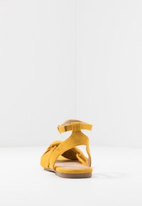 Keltainen mokkasaapas, jossa on solmittu muotoilu, avoin takaosa ja säädettävä nilkkahihna. Litteä pohja, jossa on vaalea ruskea perusta.