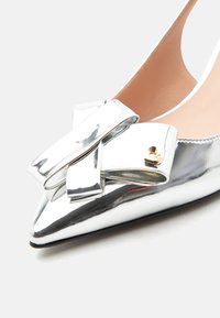 Ασημί μεταλλικό ψηλοτάκουνο με μυτερή μύτη, μεγάλο διακοσμητικό φιόγκο και μικρή χρυσή καρδιά στην περιοχή των δαχτύλων.