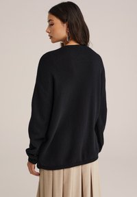 Moteris su ilgais tamsiais plaukais, vilkinti sidabrinius auskarus-žiedus, laisvą juodą megztinį ir smėlio spalvos klostuotą sijoną, pasukusi šiek tiek į šoną.