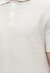 Gros plan d'une personne portant un polo blanc texturé à manches courtes avec deux boutons blancs sur la patte de boutonnage.