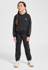 Svarta mjukisbyxor och matchande crewneck-tröja med en spräcklig textur, vita detaljer och en liten logotyp på bröstet, kombinerat med sneakers.