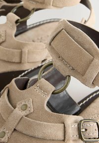 Gros plan sur les brides d'une sandale en daim beige avec des détails d'anneaux métalliques et de boucles sur une semelle foncée.