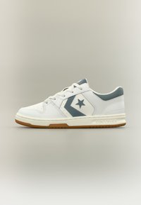 Λευκό sneaker Converse χαμηλού τύπου με γκρι αστέρι και λογότυπο σχήματος βέλους, λευκά κορδόνια και σόλα από καουτσούκ χρώματος τσίχλας σε ουδέτερο φόντο.