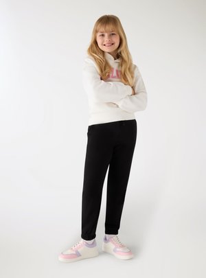 Giovane ragazza in piedi con le braccia incrociate, indossa una felpa bianca, pantaloni della tuta neri e sneakers bianche con accenti rosa e lilla.
