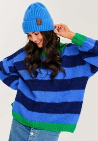 Modrý a námořnický pruhovaný oversize pletený svetr se zeleným lemem, doplněný světle modrou žebrovanou čepicí s hnědým štítkem.