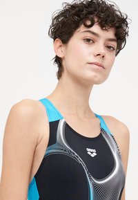 Maillot de bain noir avec des bretelles turquoises, ornée d'un motif de vagues en blanc et bleu. Le tissu semble lisse avec un léger éclat.