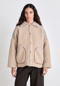 Giacca in pile beige con chiusura a bottoni, tasche ampie e colletto. Presenta accenti di tessuto a contrasto sulle spalle.