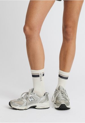 Bare ben iført hvite crew-sokker med svarte striper og sølvfargede New Balance-sneakers står på en hvit overflate.
