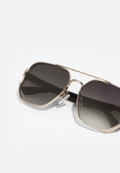 Ochelari de soare din metal aurit cu lentile maronii cu gradient, de formă pătrată, și bentițe transparente pe nas. Prezintă un design subțire cu punte dublă și brațe negre.