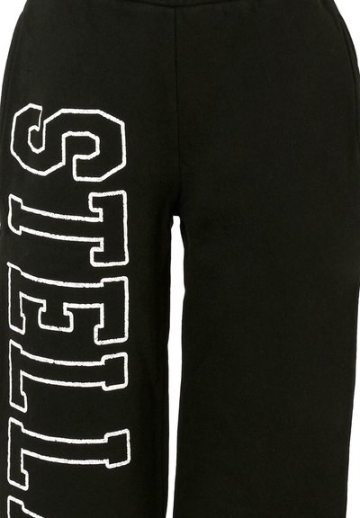 Pantalon de survêtement noir à coupe ample, avec des lettres blanches en bloc orthographiant « STELLA » le long de la jambe gauche. Texture douce, design décontracté.