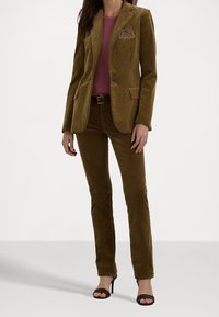 Hnědý kordový sako s vyšitým emblémem, spárované s odpovídajícími kordovými kalhotami a přiléhavým růžovým topem. Žena má na sobě černé sandály na podpatku.