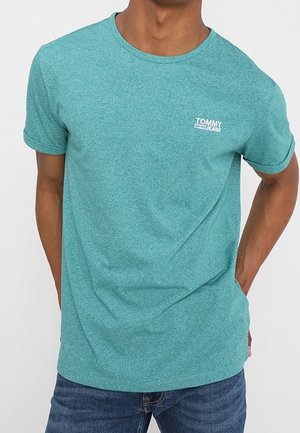 Mand iført en turkis Tommy Jeans T-shirt med op-rullede ærmer og blå jeans, stående med hænderne bag ryggen.