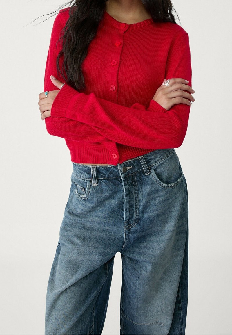 Femme portant un cardigan rouge boutonné à manches longues, les bras croisés sur la poitrine, et un jean bleu en denim ample.