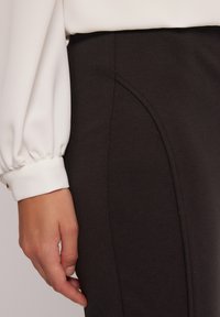 Czarne dopasowane spodnie o gładkiej strukturze, z bocznym detalem szwu. Częściowo widoczna jest biała bluzka z bufiastym mankietem.