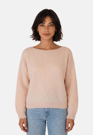 Mujer con el pelo corto y oscuro, con un jersey rosa claro y vaqueros azules, de pie frente a un fondo blanco liso.
