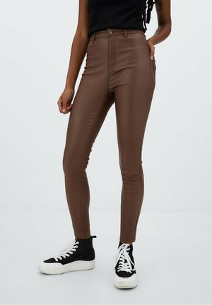 Persoon draagt hooggetailleerde bruine leren broek, zwart mouwloos topje en zwart-witte hoge sneakers, staand tegen een effen achtergrond.