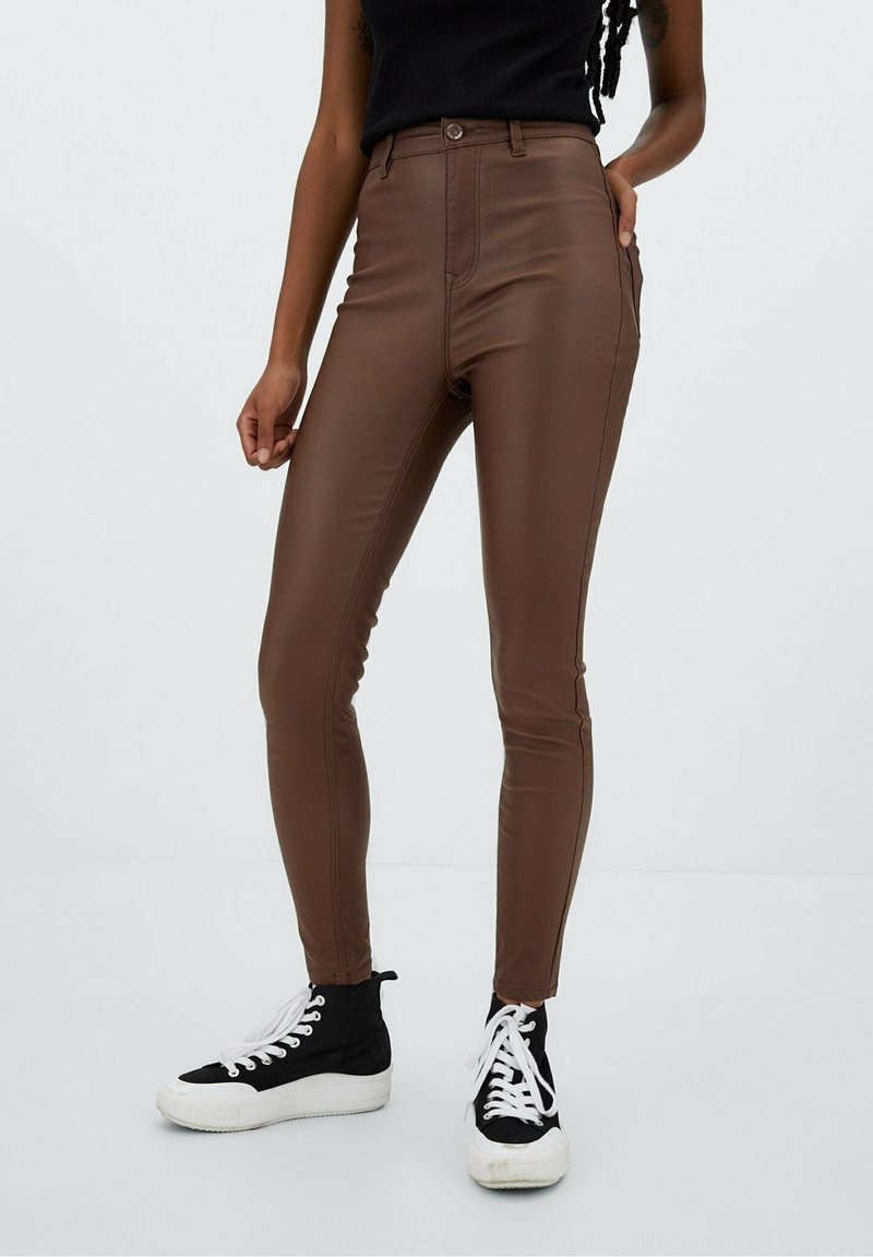 Personne portant un pantalon en cuir marron taille haute, un top noir sans manches et des baskets montantes noires et blanches, debout devant un fond uni.