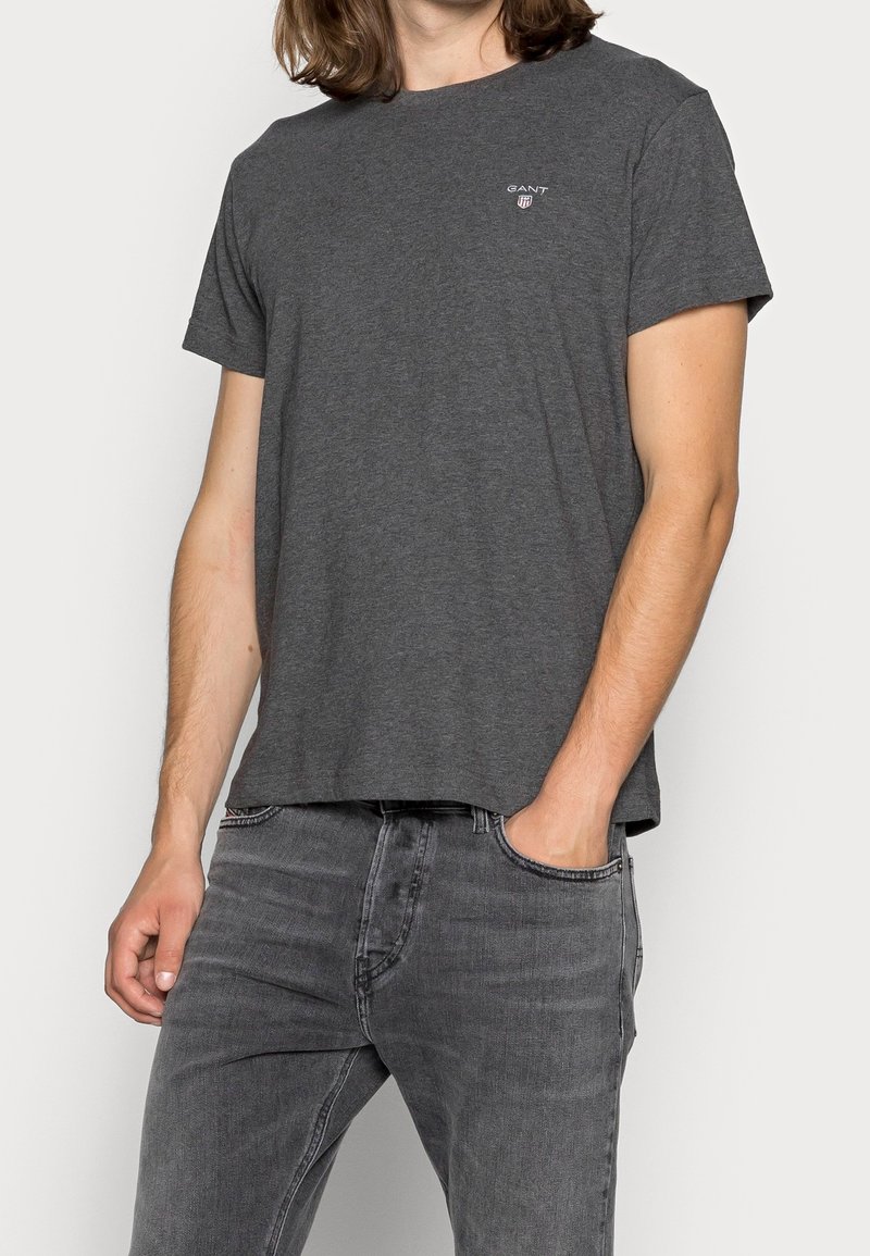 Mann trägt ein dunkelgrau GANT T-Shirt mit kurzen Ärmeln und ausgebleichten schwarzen Jeans, eine Hand in der Tasche, vor einem einfarbigen Hintergrund.