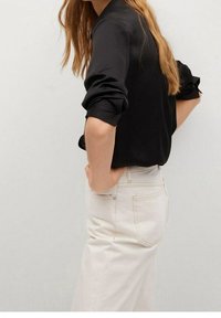 Camisa negra de manga larga con puños arremangados, combinada con jeans blancos de cintura alta, que presentan una ligera textura y un ajuste ceñido.