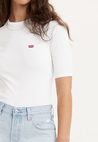 Hvid t-shirt med korte ærmer og rund halsudskæring, prydet med et lille rødt Levi's-logo på brystet, parret med lysblå slidte denimjeans.