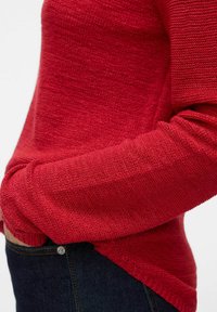Rdeča pletena sv sweater z teksturiranim finišem. Ima sproščen kroj, okroglo ovratnico in rebraste manšete. Nosi se preko temno modrih džinsov.