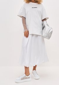 T-shirt bianco oversize con logo "JIL SANDER", abbinato a una gonna plissettata bianca e sneaker argento con un motivo testurizzato. Borsa argento in risalto.