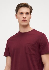 Burgundy short-sleeve t-shirt with a crew neck and a left chest pocket, made of soft fabric with a smooth texture.