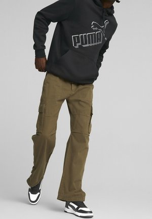 Osoba ubrana w czarną bluzę z kapturem marki Puma, oliwkowe spodnie typu cargo i czarno-białe trampki, stojąca na tle jednolitego szarego tła.