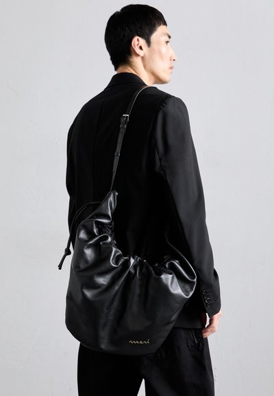 Vyras juodame kostiume laiko didelę juodą odinę petinę krepšį su raukšlėtuvu viršumi ir mažu auksiniu logotipu prieš pilką sieną.