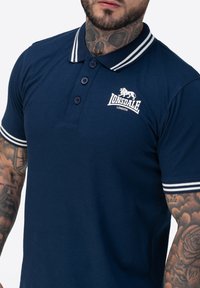 Navyblaues Polohemd mit weißem Streifenkragen und -manschetten, einer Dreiknopfleiste und einem kleinen Lonsdale-Logo auf der Brust.