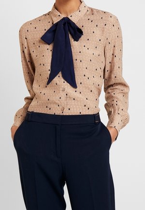 Beige mönstrad blus med en marinblå rosettkrage instoppad i marinblå byxor med hög midja, händerna i fickorna.