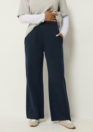 Personne portant un pantalon large bleu marine, des hauts gris superposés et des baskets blanches, debout avec une main dans la poche et l'autre sur le ventre.