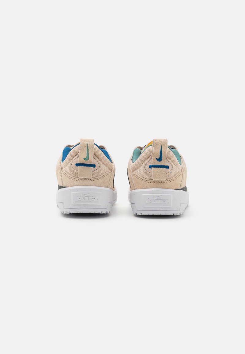 Beige Wildlederschuhe mit weißen Gummisohlen, die blaue und grüne Akzente an der Ferse und Zunge aufweisen, mit einer Zuglasche an der Rückseite.