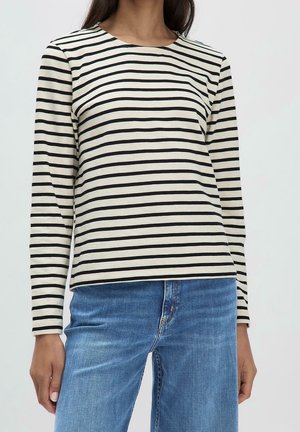 Mujer con una camisa de manga larga color crema con rayas horizontales negras, combinada con jeans de mezclilla azul, posando frente a un fondo sencillo.