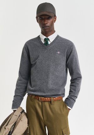 Mann trägt einen grauen V-Ausschnittpullover, weißes Hemd, grün gestreifte Krawatte, braunen geflochtenen Gürtel, olivfarbene Hose, schwarze Mütze, hält eine beige Jacke in der linken Hand.