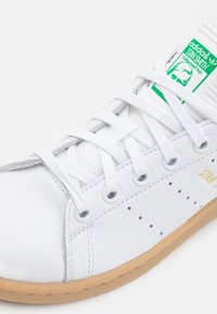 Bílé kožené tenisky s gumovou podrážkou, s šněrovacím designem, perforovanými detaily a zeleným štítkem s logem na jazyku.