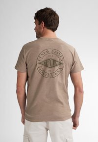 Beige lyhythihainen t-paita pehmeästä kankaasta, jossa on iso pyöreä vihreä logo selässä ja teksti "DENIM DRIVEN PETROLIND."