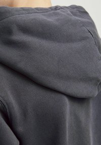 Felpa grigia che mostra un tessuto texture con cappuccio regolabile e cuciture visibili, appoggiata su uno sfondo neutro.