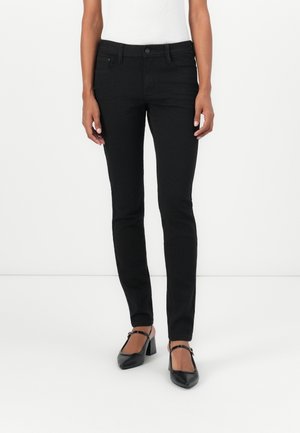 Nainen pukeutunut mustiin slim fit -farkkuihin, mustiin Mary Jane -korkokenkiin ja valkoiseen hihattomaan toppiin, seisoo valkoista taustaa vasten.