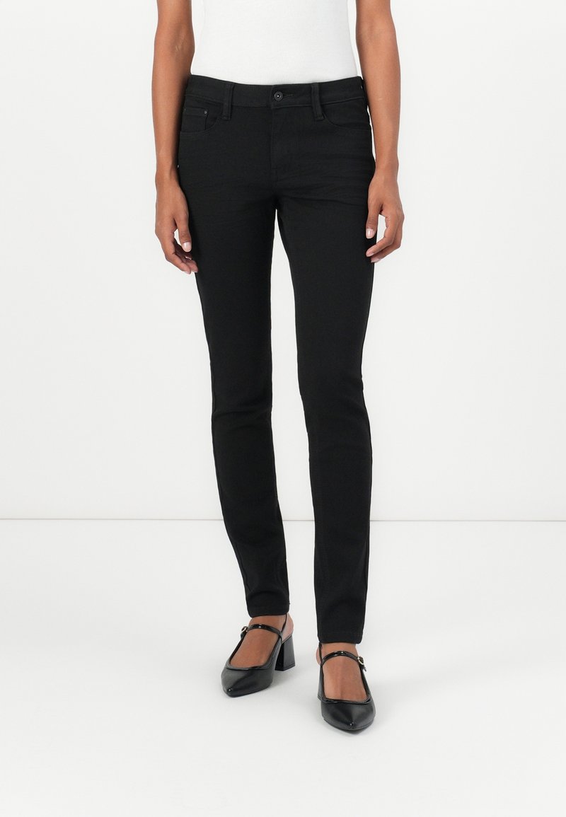 Femme portant un jean noir slim, associé à des talons Mary Jane noirs et un top blanc sans manches, debout sur un fond blanc.