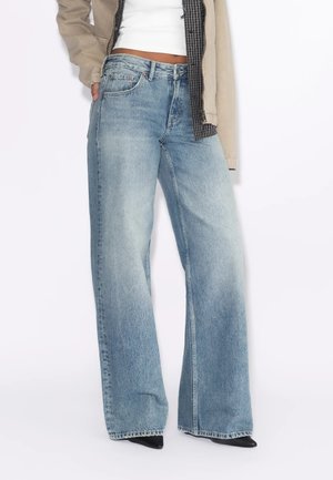 Person trägt hellblaue Jeans mit weitem Bein, schwarze spitze Schuhe, weißes Oberteil und beige Jacke mit kariertem Futter, steht vor einem weißen Hintergrund.