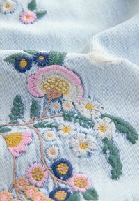 Broderie de fleurs colorées et de feuilles vertes cousues sur un tissu en denim bleu clair avec une petite déchirure effilochée près du motif.